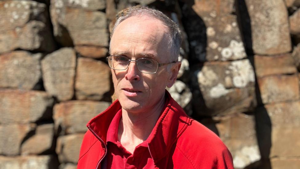 Dr Henry is looking at the camera and is wearing thin framed glasses. He has thin grey hair and is wearing a bright red fleece and polo top. 