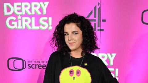 Jamie Lee- O'Donnell has long brown bushy hair and is wearing a black T-shirt with a yellow smiley face. She is standing in front of a pink board with a number of signs.