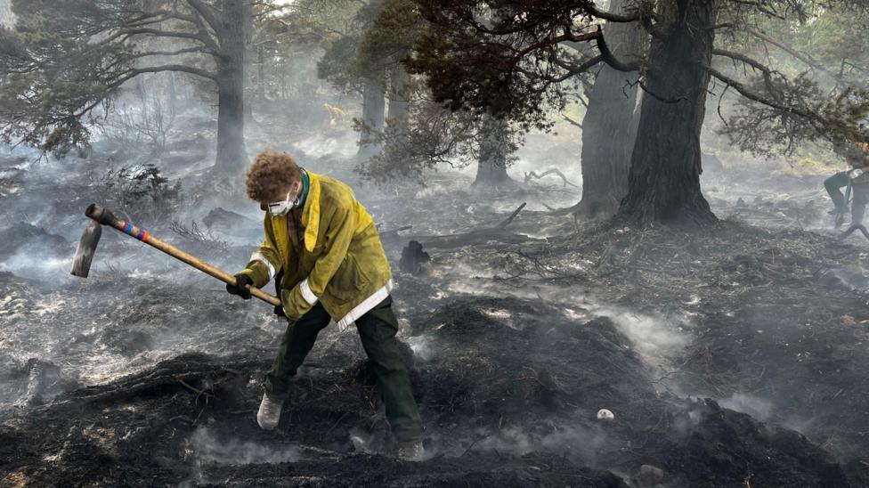 A person wearing a yellow protective coat and a face covering uses a pick to break up burning peat and vegetation at Abernethy Nature Reserve.