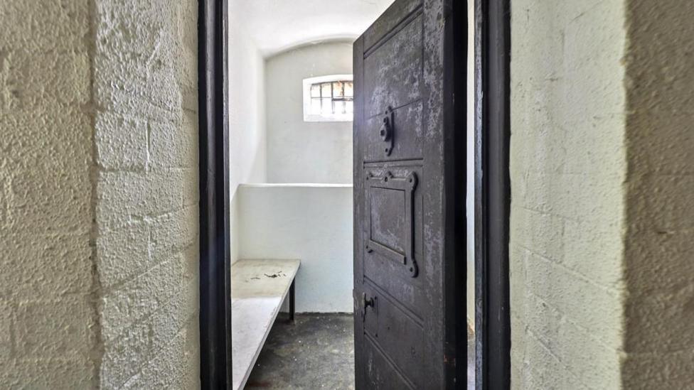 An imposing heavy grey metal cell door stands ajar, a bed can just be seen inside.