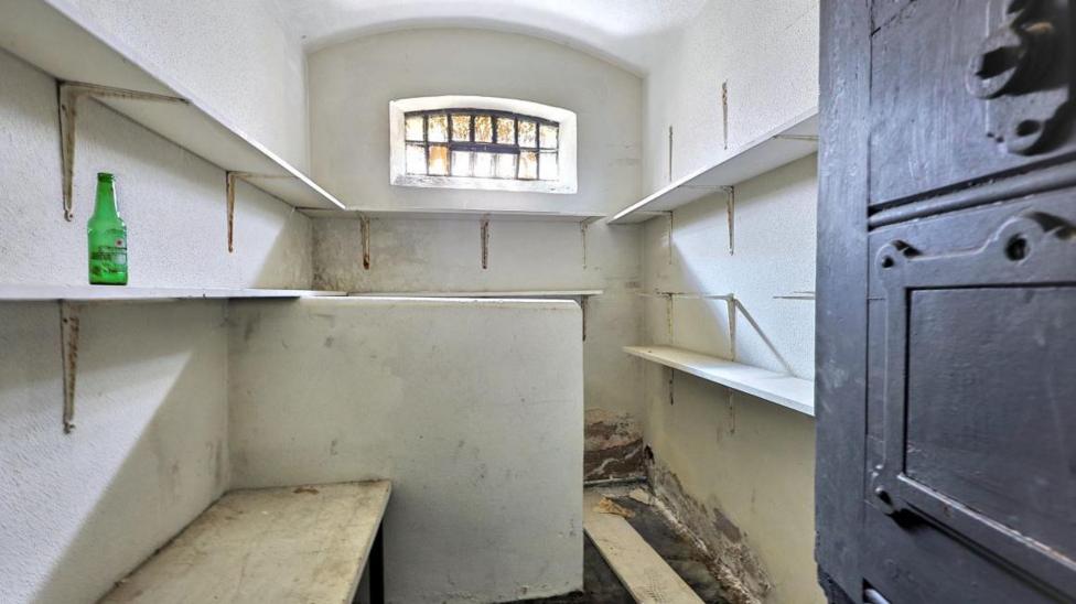 Inside view of one of the cells, there's a hard wooden bench on one side and a low wall beyond it obscuring the other side. Shelves line the wall, with a green glass bottle on one of them.