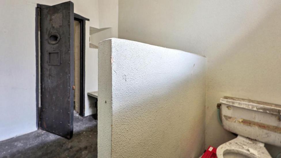 Inside view of one of the cells, showing a white toilet obscured by a low wall with a cell door beyond it