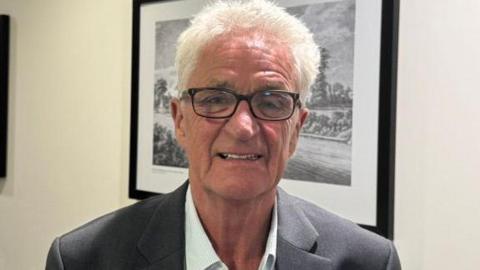 A man wearing a grey suit adn white shirt. He has black glasses and grey hair and is stand in front of a black and white photo in a frame.