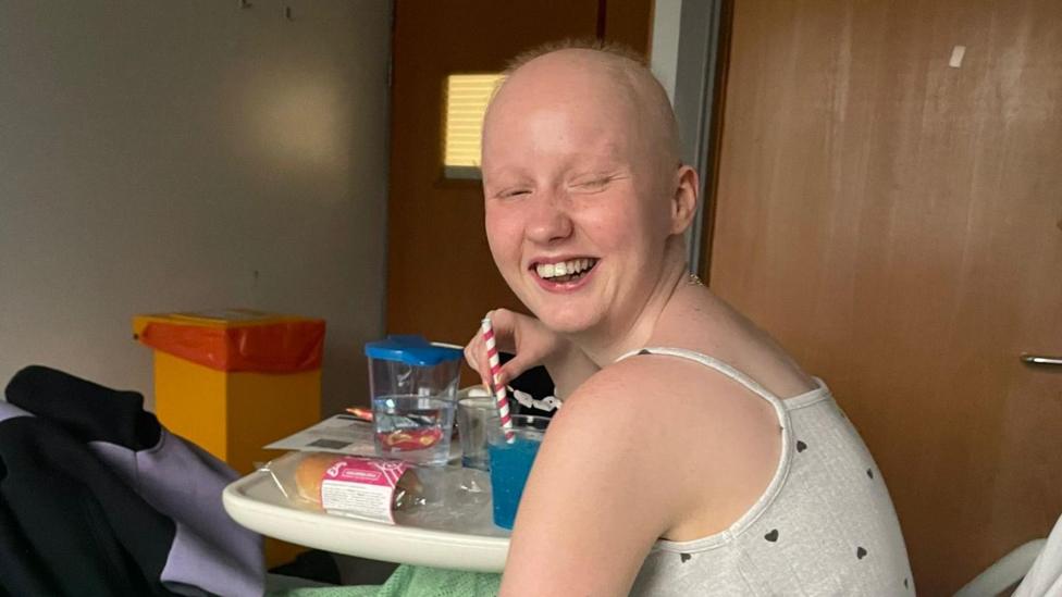Enlli Thompson is photographed in a hospital bed. She is wearing a grey vest top which is covered in a pattern of small black hearts. She is drinking a blue slush drink and holding a white and red straw. She looks at the camera, smiling and winking.