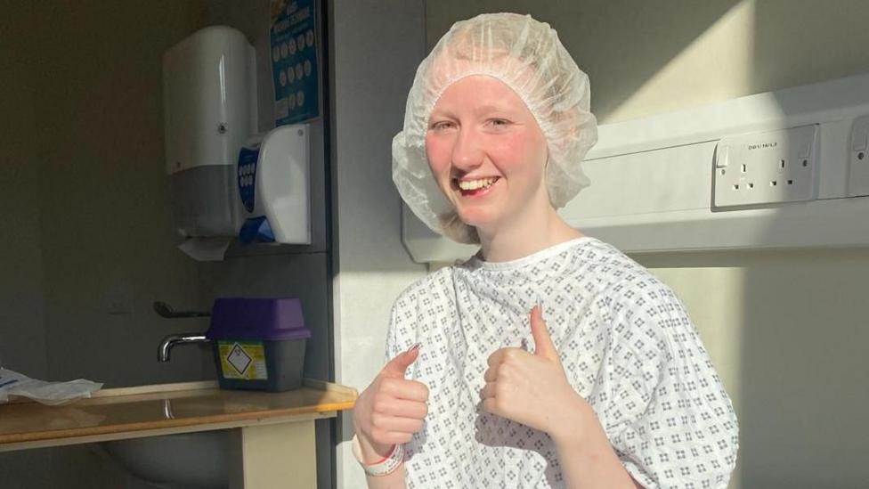 Enlli Thompson is photographed in hospital. She is smiling, looking at the camera with both thumbs up. She is wearing a hair net and a hospital gown.