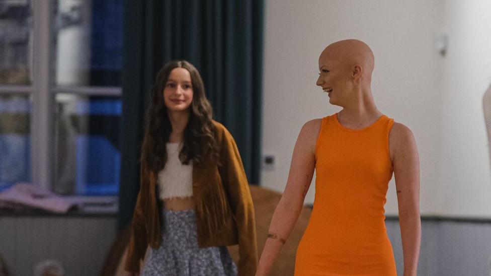Enlli Thompson taking part in a fashion show for the charity, Tenovus Cancer Care. Her head is shaven and she is wearing a short sleeve orange dress. To her right is another young girl who is also taking part in the fashion show. She is wearing a brown jacket, white crop top and a purple floral skirt.