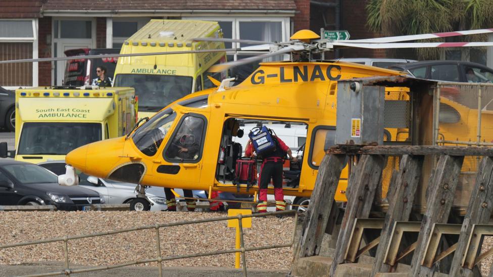 An air ambulance helicopter is parked on the beach near Shoreham Port with ambulance vehicles and emergency personnel nearby.