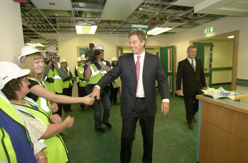 A man in a suit with a white shirt and pink tie is smiling and shaking the hands of a row of workers in hard hats and hi viz vests