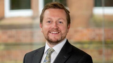 A man smiling at the camera wearing a suit and tie. The jacket is black and a white shirt with a floral tie. He has ginger hair and a beard.