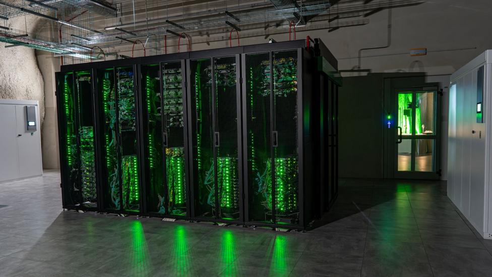 Green lights shine from a row of data servers in cabinets, reflecting on the high-gloss floor of the underground cavern. Metal trunking is attached to the ceiling above the cabinets, while to the right a door leads into a lighter room.