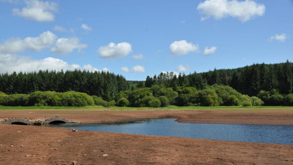 A dried out reservoir bed with a small amount of blue water and an old stone bridge. Green grass, trees and blue sky with clouds are in the background.
