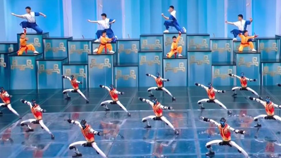 A screengrab shows robots in gold and red vests performing martial arts on stage during a Lunar New Year showcase, alongside young athletes carrying out a similar routine in the background.