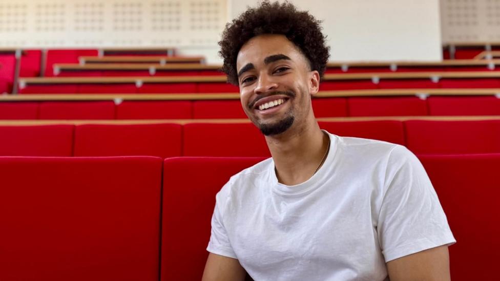 Kieran McRae is in a white T-shirt seated in a lecture theatre with rows of bright red tiered seats. The image is framed as a portrait, with the seating stretching into the background of the auditorium.