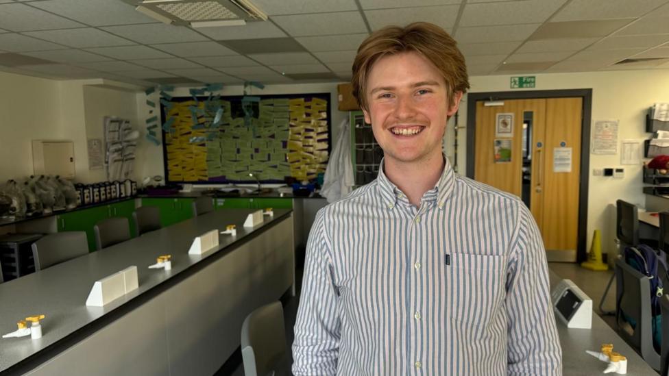 Daniel is seen standing in a science classroom or laboratory, wearing a striped shirt. Long workbenches with taps and stools fill the room, while a noticeboard covered with coloured papers and a door marked with a fire exit sign are visible in the background.