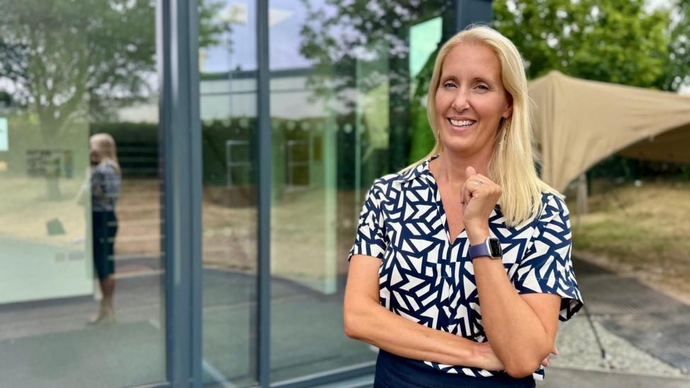 Vicky Orsmond is seen standing outside a modern glass-fronted building, wearing a patterned short-sleeved shirt and a smartwatch. A reflection is visible in the window behind, with trees, a canopy structure and landscaped grounds in the background.
