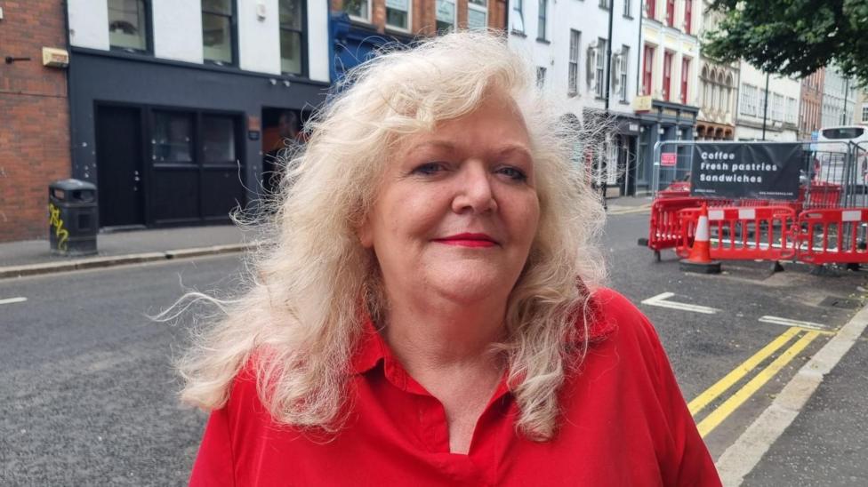 A woman, who has long light blonde hair, wearing a red shirt. She is standing in front of a road in Belfast city centre.