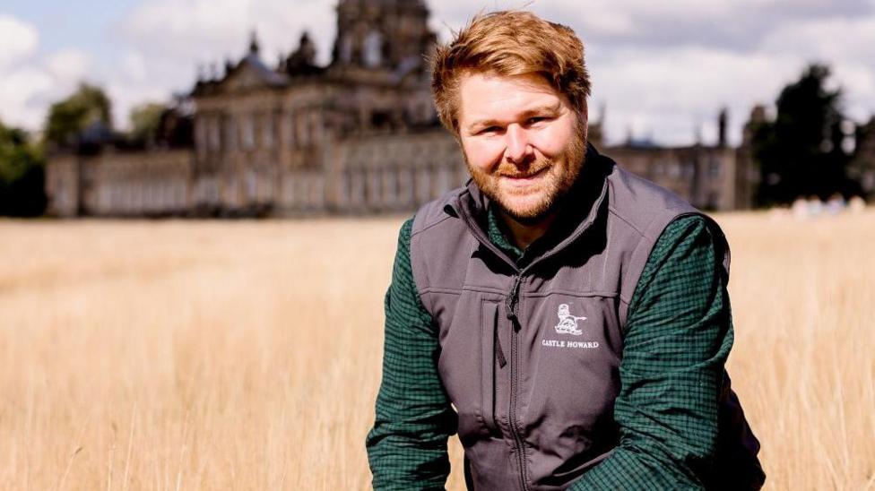 A man, Guy Thallon, wears a green and navy chequered shirt and a Castle Howard branded gilet. He is kneeling in a field with the large historic house in the background.