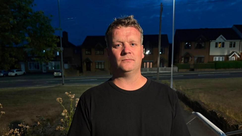 A man in a black t-shirt with grey hair, he is looking towards the camera with a stern look.