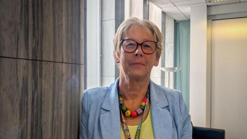 A woman with short blonde hair is wearing a blue blazer and a yellow blouse. She is looking towards the camera with a slight smile.