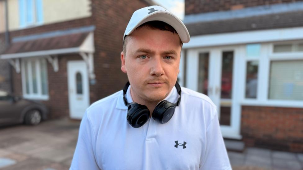 A man with a white cap and polo shirt on, he has black headphones round his neck and looking towards the camera.