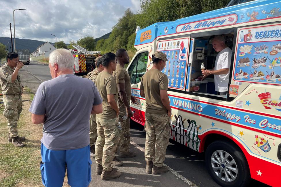 A group of soldiers are stood in a queue towards an ice cream van. In the van is a man holding out an ice cream.