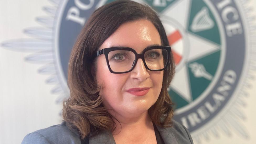 A woman with black glasses wearing a grey blazer. She has brown hair.