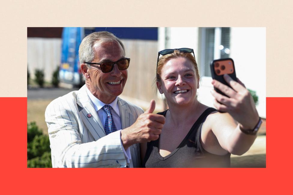 Reform UK party leader Nigel Farage poses for a selfie with a woman on the day of the Clacton by-election. He is wearing sunglasses, a blue and white striped blazer, a white shirt and blue tie, and is giving a thumbs up gesture. The woman holds the phone with her outstretched arm.
