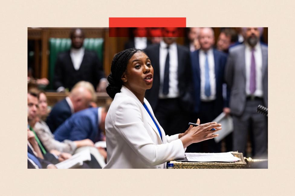Kemi Badenoch, viewed in profile, addresses the House of Commons from the despatch box. She wears a white jacket, a blue top and is holding a pen. The blurred outlines of other MPs are visible in the background.