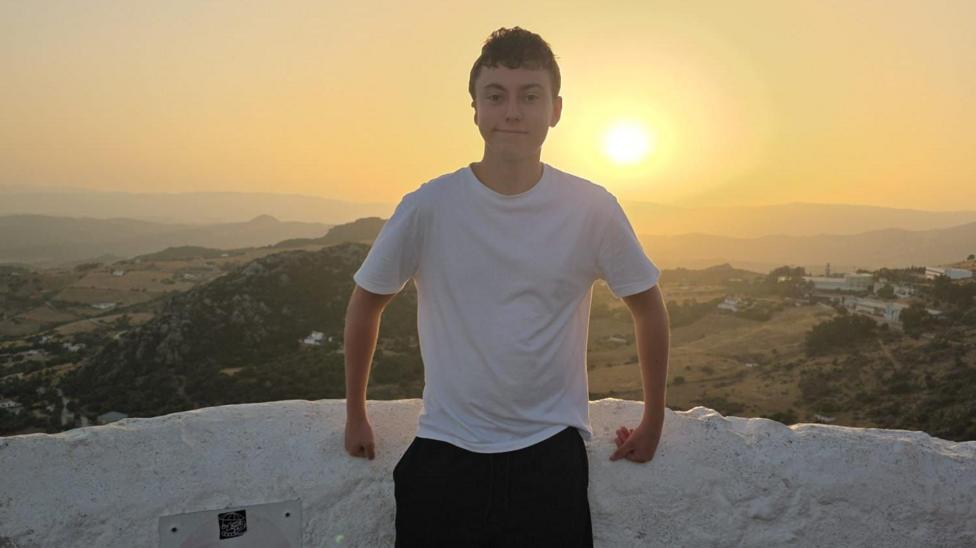 Will Payne smiles at the camera. He is stood in from of a sunset and hills. he is wearing 