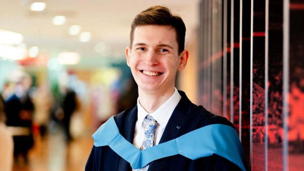 Rainers is wearing a dark blaze, white shirts ad colourful tie, and across his chest and draped from his shoulders is a blue piece of material traditionally worn by people on graduation day.