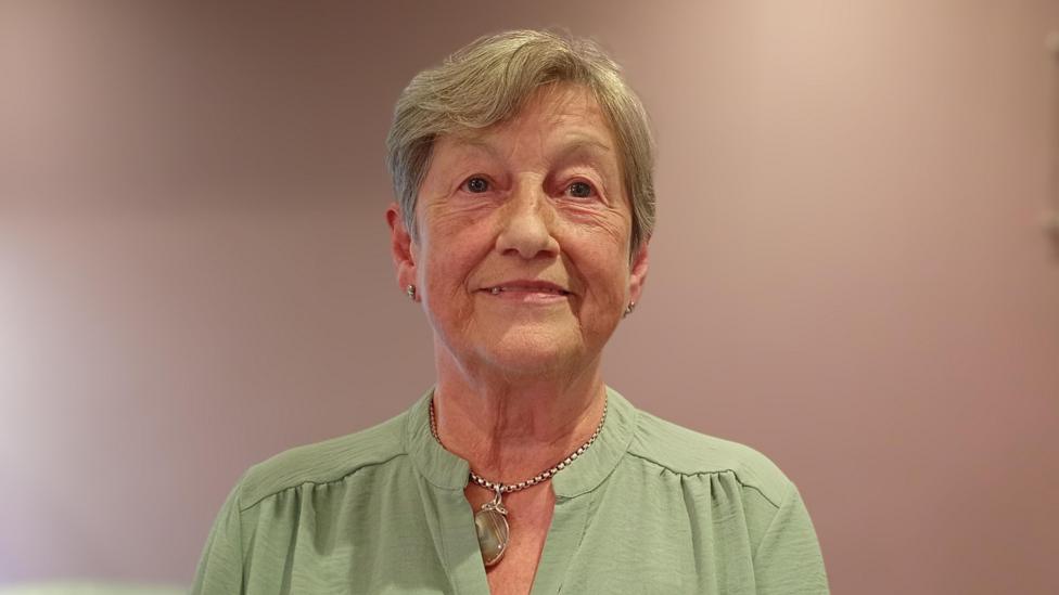 A woman, with short grey hair, wearing a green top and a necklace.