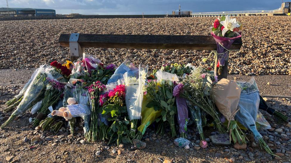 A pile of flowers left by a wooden post next to a shingle beach.