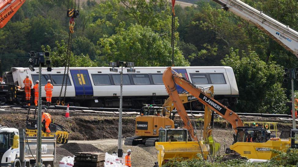 Heavy machinery including cranes, excavators and construction vehicles is being used at the train derailment site near Lewes. A Southern train carriage is positioned beside the track while recovery crews work around the railway embankment and construction materials.