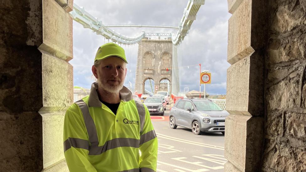 A middle-aged man wearing yellow hi-vis on a large bridge crossing. There's cars travelling in both directions.