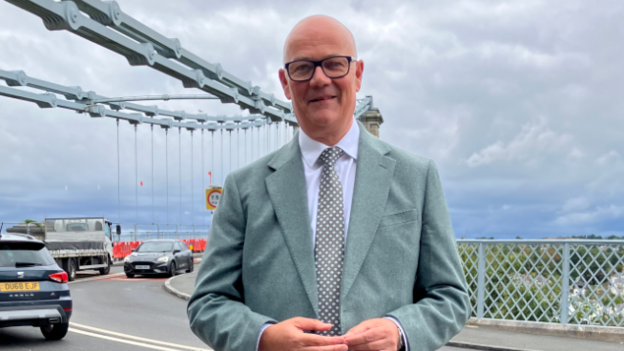 A middle-aged bald man in a grey suit stood outside a large bridge crossing with cars travelling in both directions.