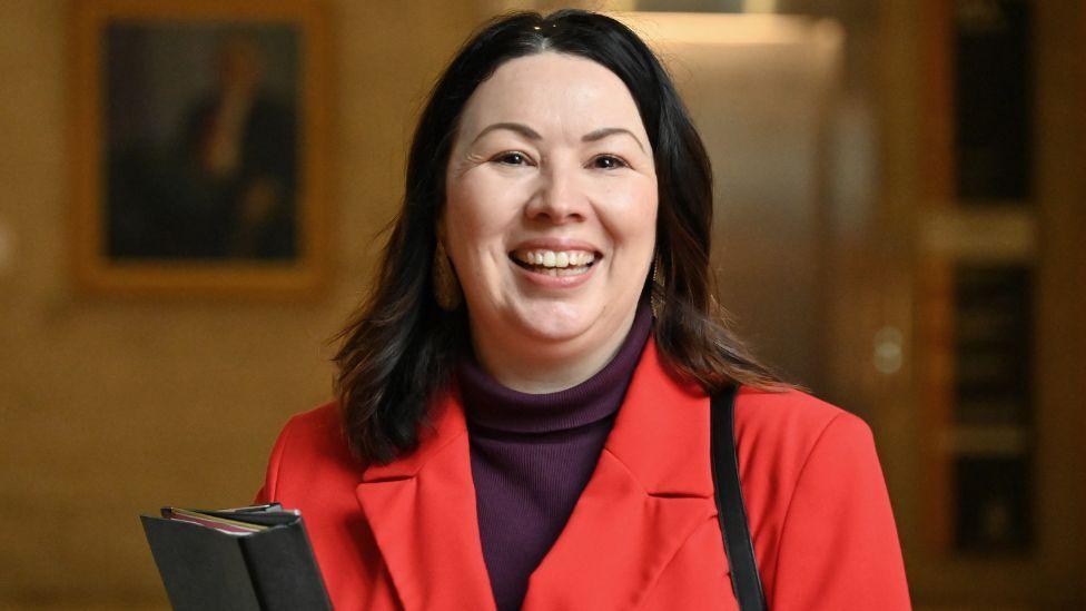 Monica Lennon, who has shoulder-length dark hair, smiles while looking at the camera. She is wearing a red jacket and carrying a black folder