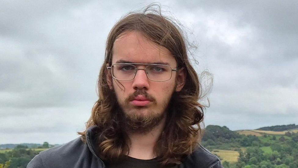 Tobias has long hair and glasses and is standing outdoors in front of rolling countryside under a cloudy, overcast sky.