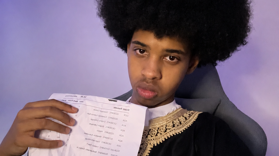 Adbi, with dark afro-style hair holds a crumpled printed document and wears a dark tunic with gold embroidered trimgainst a wall lit by blue LED lights.