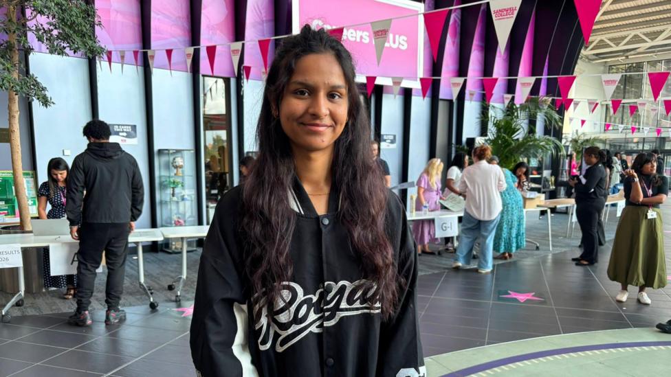 Amandeep stands in a college atrium wearing a black varsity-style jacket, with registration tables, pink bunting, and groups of people in the background