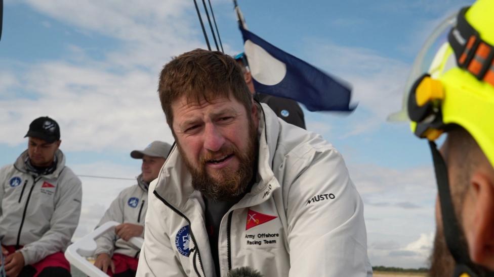 Three crew members wearing white jackets sit on a boat. There is a blue flag flying in the background.
