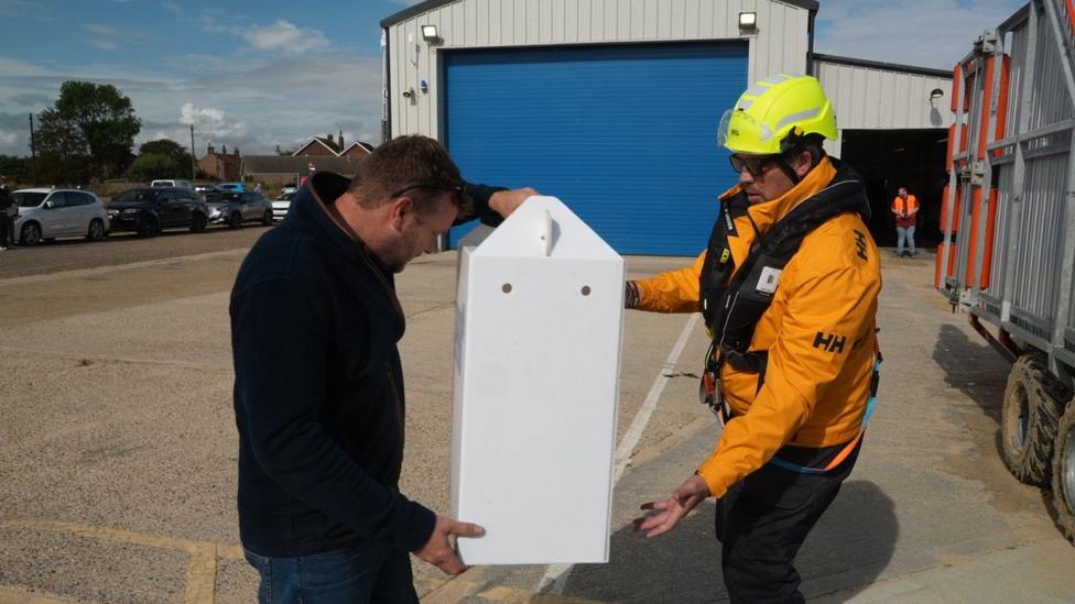 A man in a life vest and yellow jacket and neon yellow helmet hands over a tall white box, containing the owl, to a man in a black fleece and jeans.
