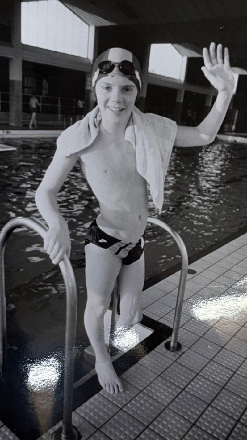 A black and white photo of Mark as a child, waving at the camera from the steps of a swimming pool. He is wearing swimming trunks and a swimming cap, and his left leg is amputated below the knee.
