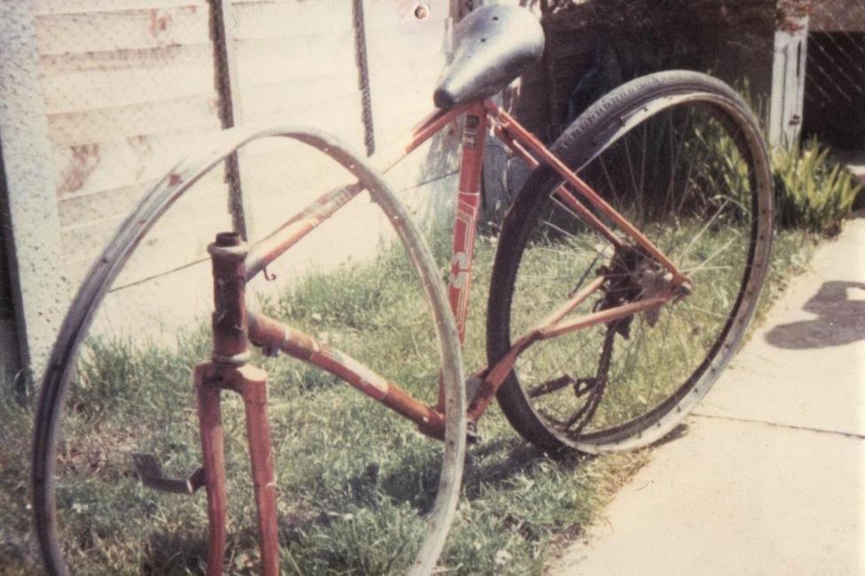 An old photo from 1982 of Mark Williams' damaged bicycle. It is a red road bike with a slightly damaged back wheel and the chain broken. The front wheel is missing, and the metal wheel rim with no spokes is leaning up against the frame of the bike.