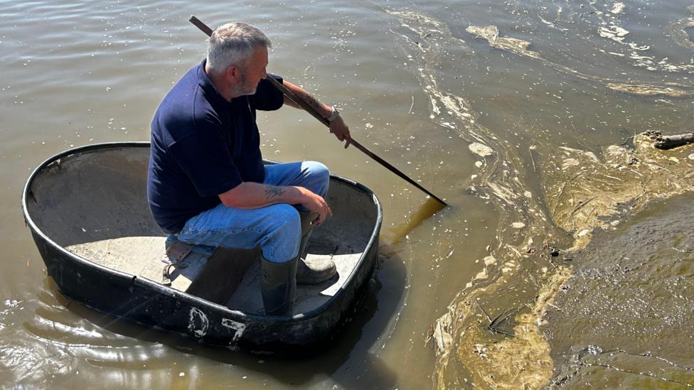 Andrew Davies, grey hair and wearing a blue polo shirt, blue jeans and green wellies. He's sitting in a coracle; a small round boat holding his oar in the water. He's looking at dirty water with lots of mud around him.
