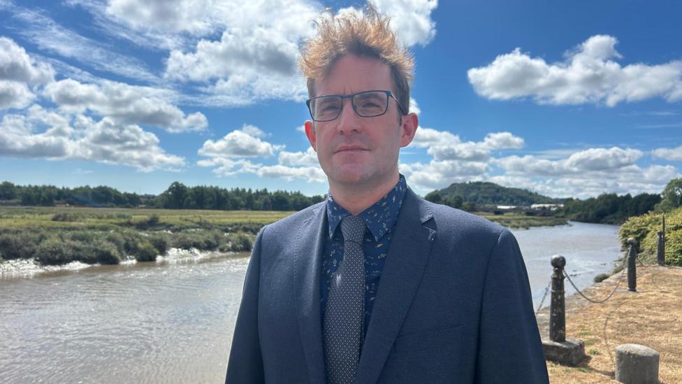 Russell Sparks in a pattered shirt, blue tie and navy jacket. He's wearing glasses and has dark hair. He's standing on the banks of the river Tywi.