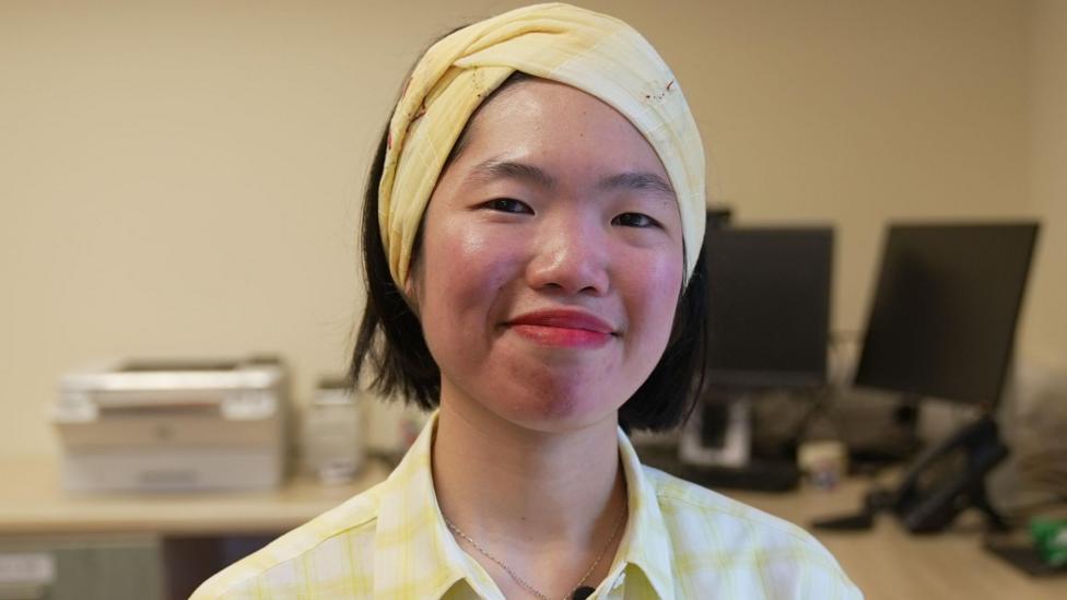 Dr Gillian Chin is wearing a checked yellow shirt and headband. She is in her GP surgery - Erskine Medical Practice in Dundee