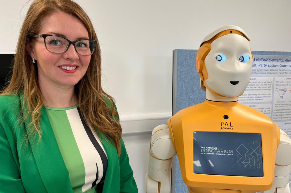 Lisa Farrell is wearing a green top and jacket and stands beside a humanoid robot at the National Robotarium