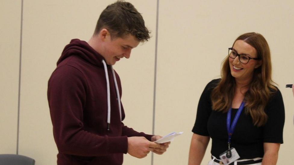 A young man in a maroon hooded top is looking at a sheet of paper that contains his exam results. He is smiling. A woman with dark hair and glasses in a dark top is also smiling.