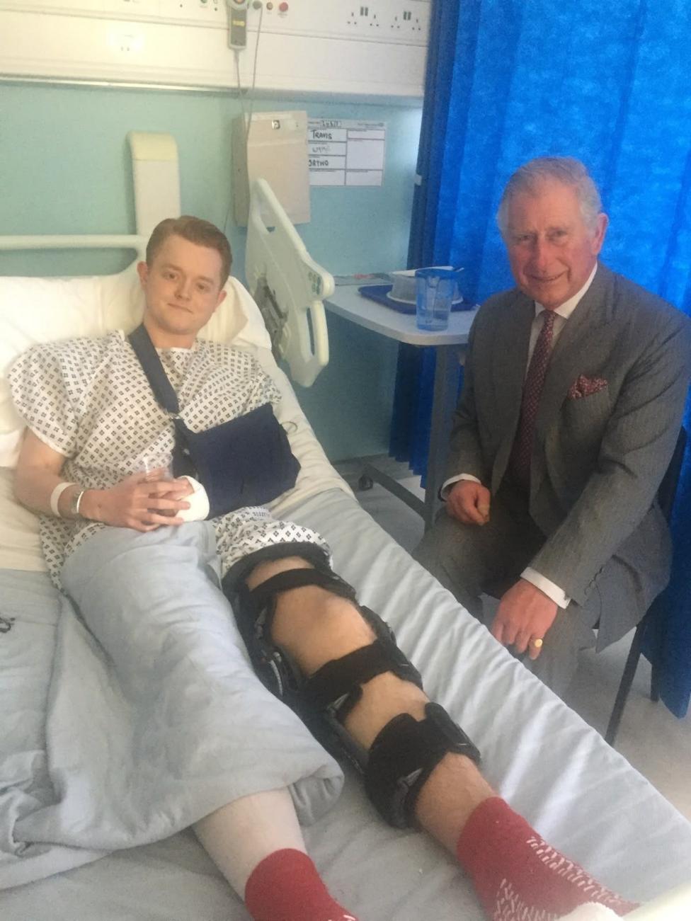 Travis Frain lays on a hospital bed with his leg strapped while King Charles is at his bedside.