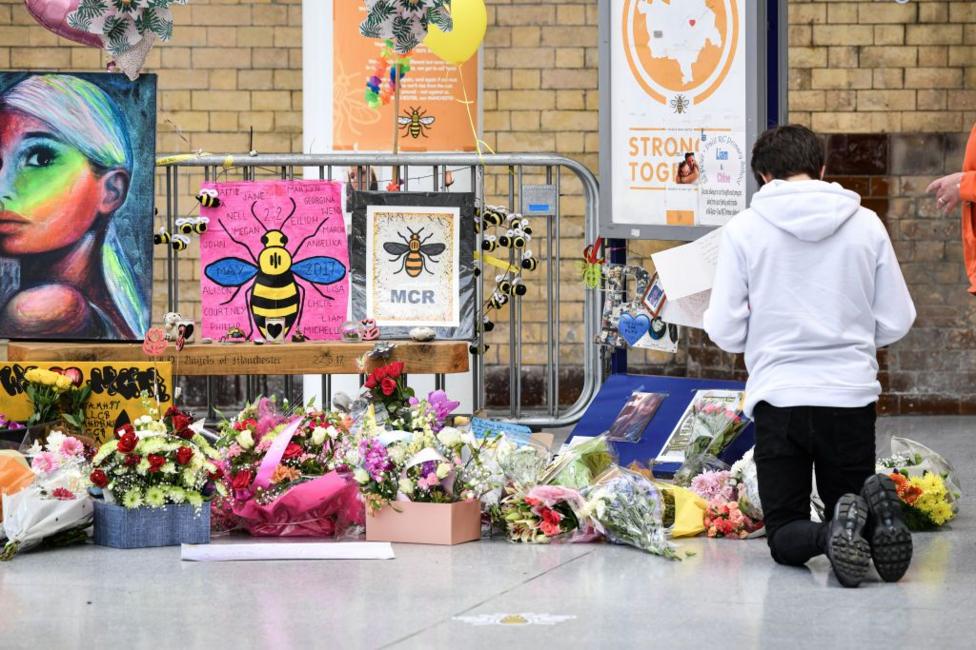 A young man kneels in front of the floral memorial at Victoria station during the commemoration. Colourful flowers, posters and pictures of Manchester bee icons are shown.